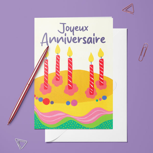 Carte pliée Bougies d'Anniversaire posée avec un stylo doré et son enveloppe, gâteau garni de bougies allumées