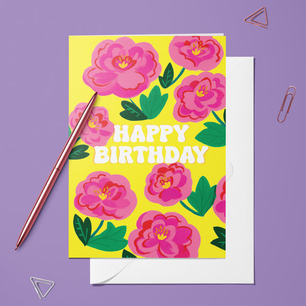 Carte pliée Pétal Party posée avec un stylo doré et son enveloppe, message d'anniversaire sur un fond de pivoines roses