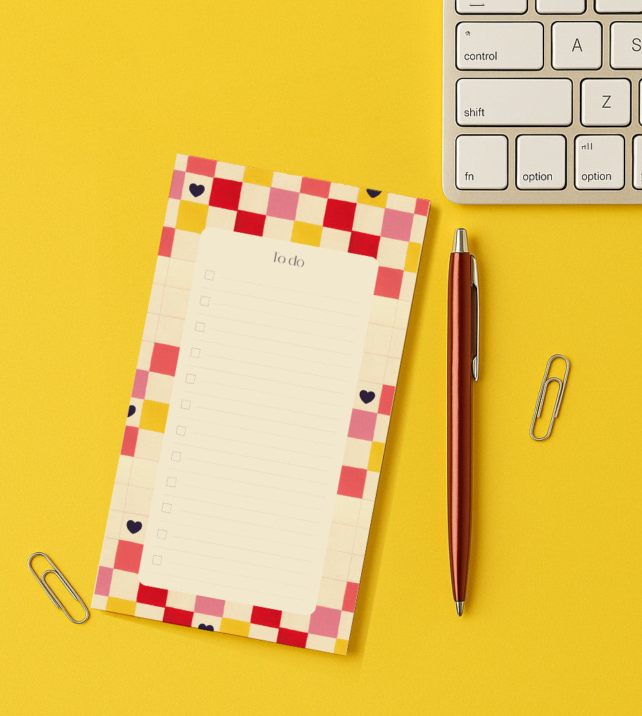 To-do Damier cœur posée sur un bureau jaune, liste encadrée d'un damier rouge et rose, près d'un stylo