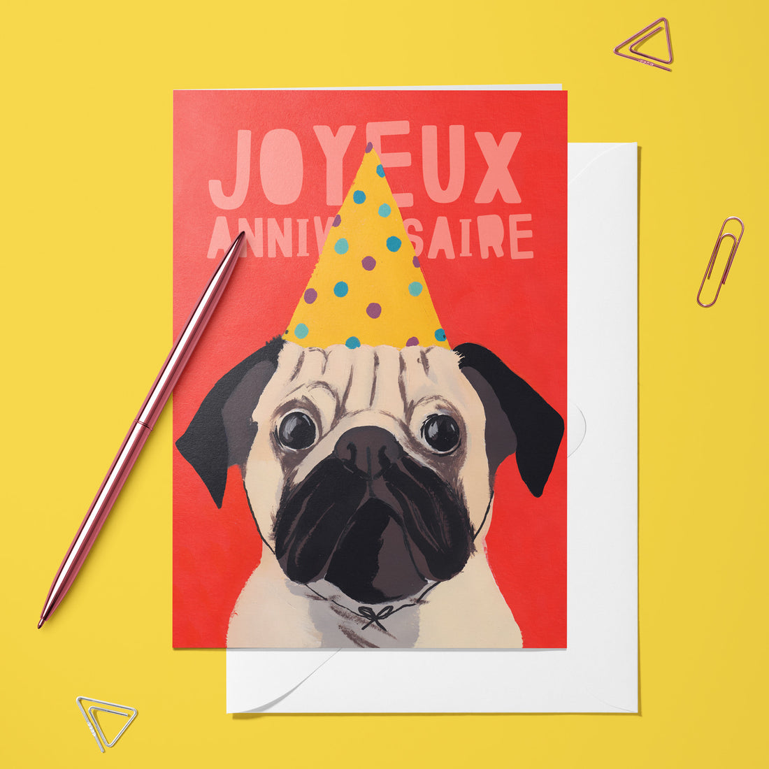 Carte pliée Bouledogue anniversaire posée avec un stylo doré et son enveloppe, carlin coiffé d'un chapeau de fête