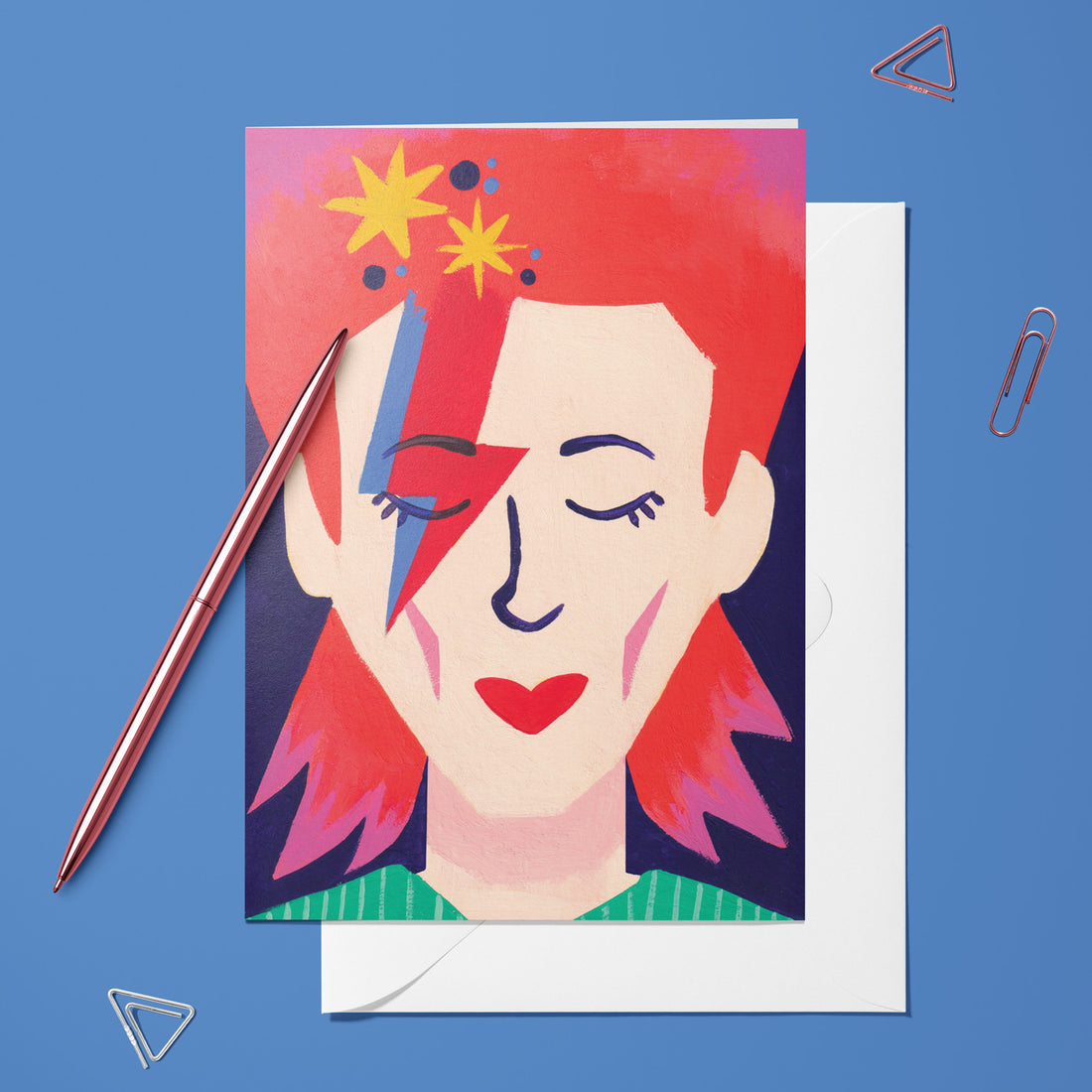 Carte pliée Bowie posée avec un stylo doré et son enveloppe sur un fond bleu