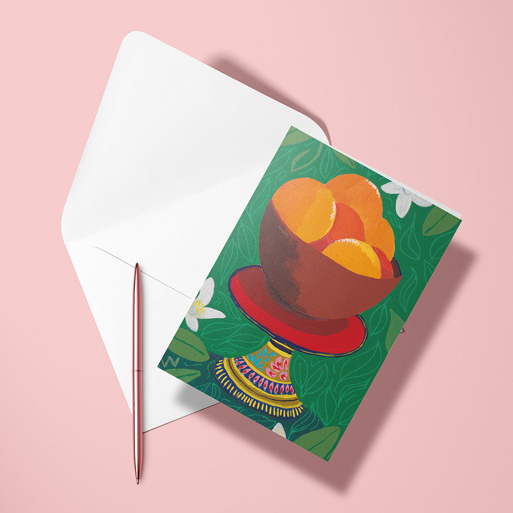 Carte pliée Coupelle d'Oranges posée avec un stylo doré et son enveloppe sur un fond rose
