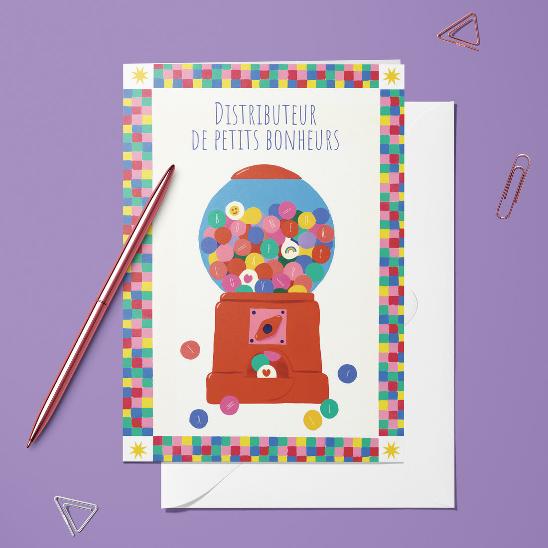 Carte pliée Distributeur de petits bonheurs posée avec un stylo doré et son enveloppe, distributeur rempli de boules colorées