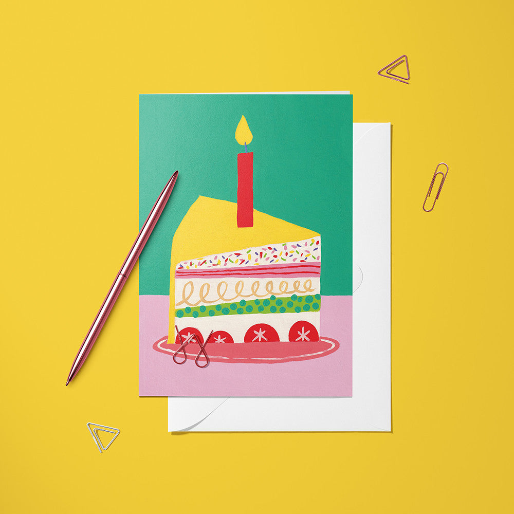 Carte pliée Gâteau d'Anniversaire posée avec un stylo doré et son enveloppe, part de gâteau et bougie allumée
