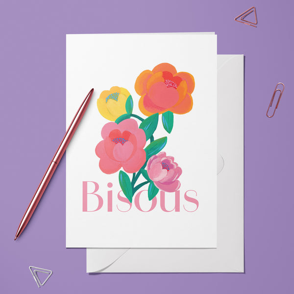 Carte pliée Bisous Éclat de Pivoine posée avec un stylo doré et son enveloppe sur fond violet
