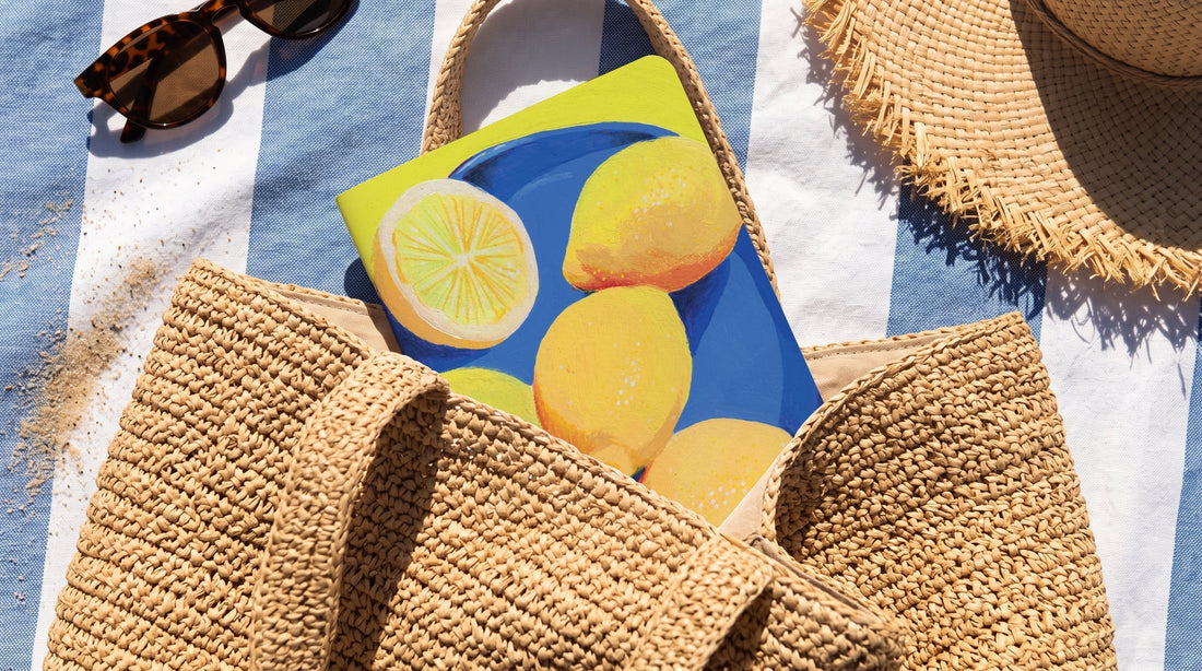 Carnet illustré Hello couleur à motif de citrons, glissé dans un sac de plage en paille sur une serviette rayée