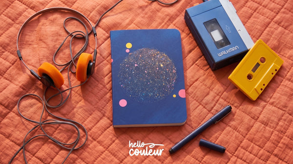 Carnet illustré Hello couleur bleu nuit à motif pailleté, posé sur un plaid orange avec un casque audio et un walkman vintage