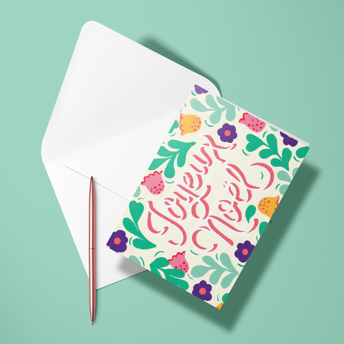 Carte pliée Joyeux Noël posée avec un stylo doré et son enveloppe, message calligraphié entouré de feuillages