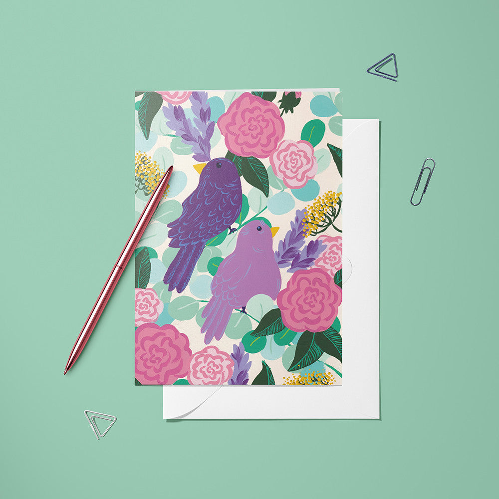Carte pliée Oiseaux posée avec un stylo doré et son enveloppe, oiseau violet perché parmi les roses