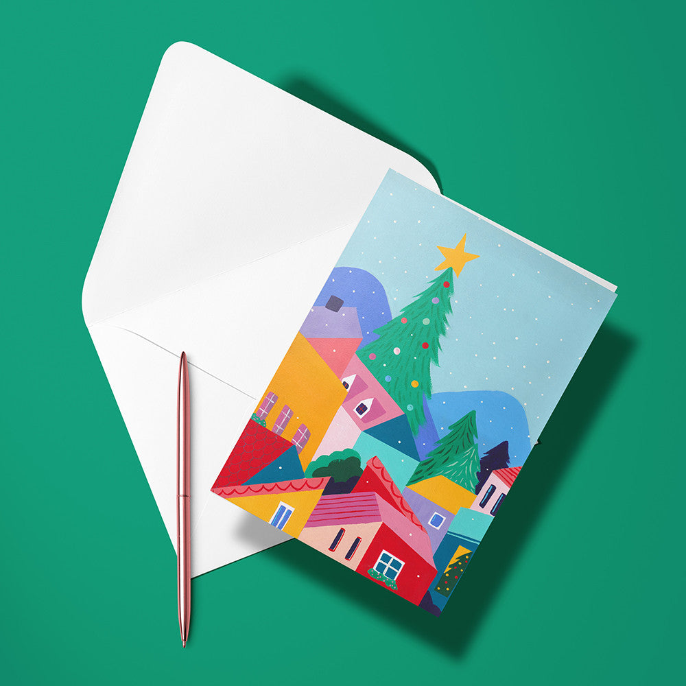 Carte pliée Village de Noël fleuri posée avec un stylo doré et son enveloppe, village coloré et sapin décoré