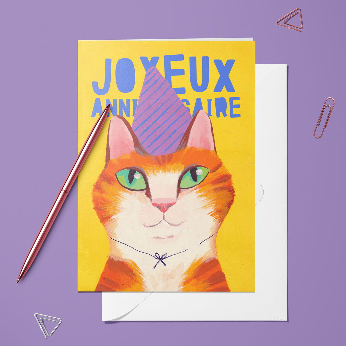 Carte pliée Chat Anniversaire posée avec un stylo doré et son enveloppe, chat roux coiffé d'un chapeau de fête
