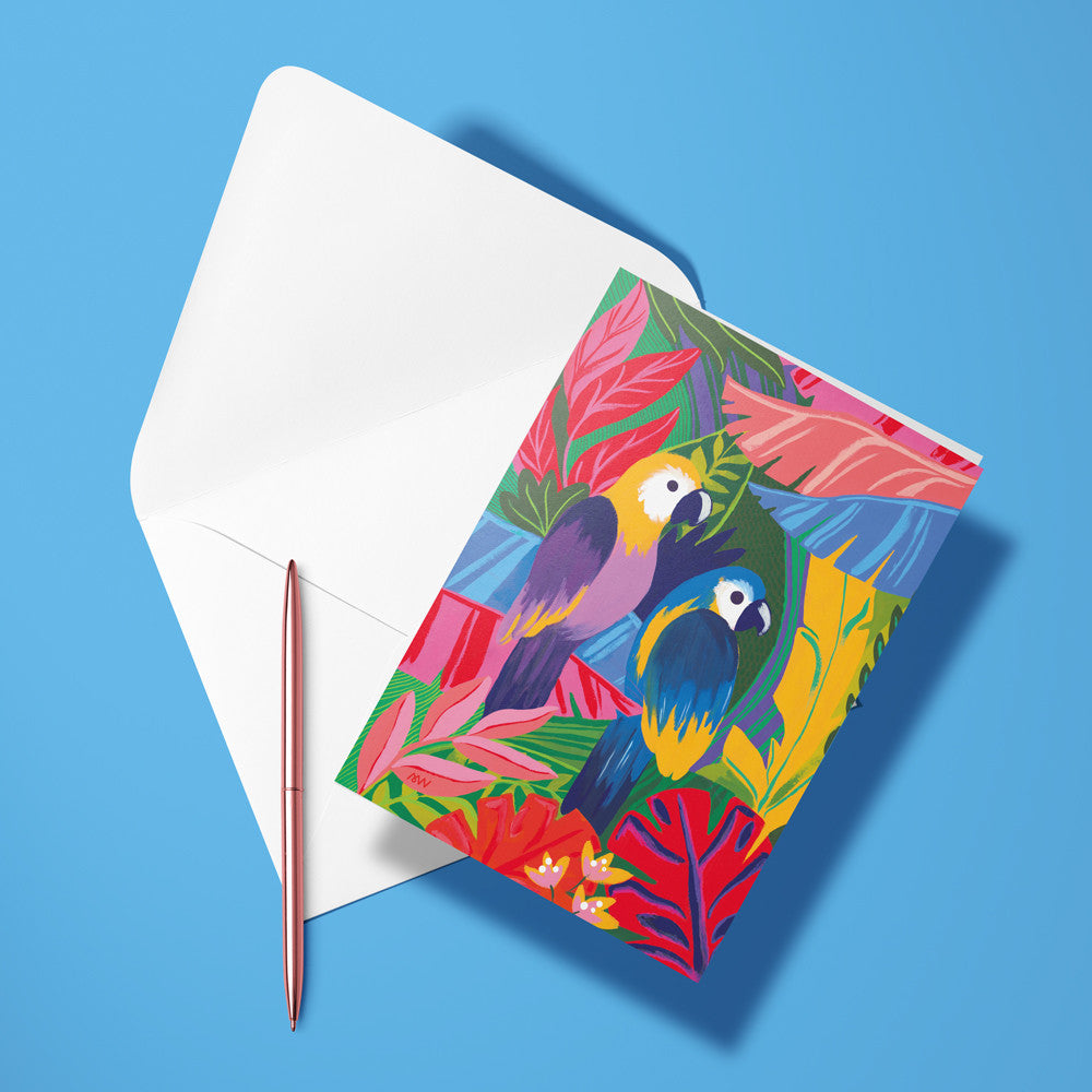 Carte pliée Les Perroquets posée avec un stylo doré et son enveloppe, deux aras dans un feuillage tropical