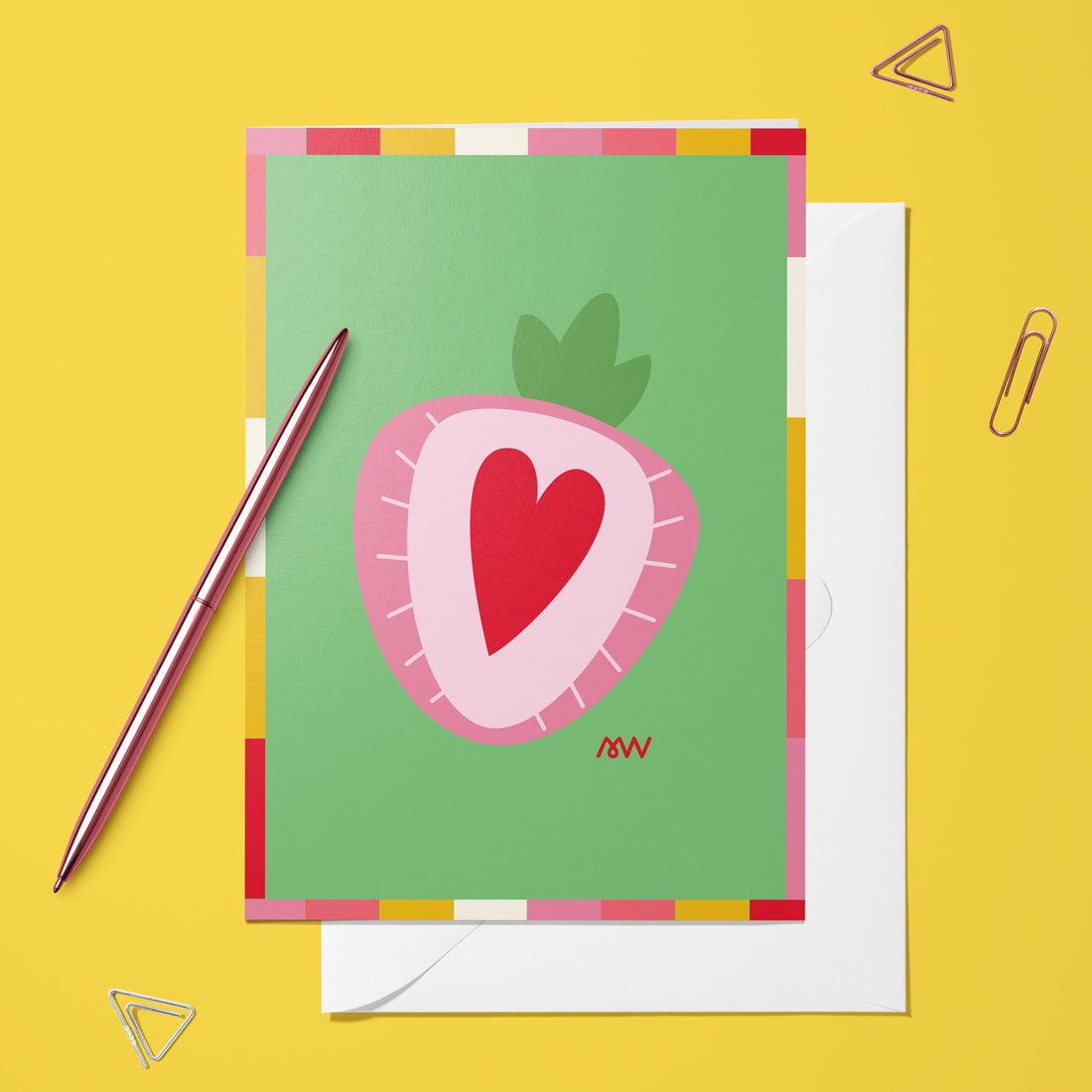 Carte pliée Cœur de Fraise posée avec un stylo doré et son enveloppe sur un fond jaune