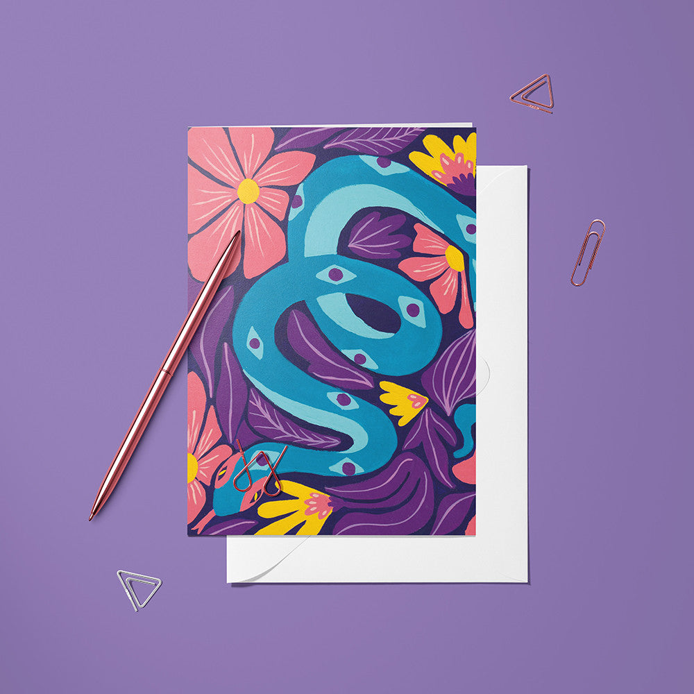 Carte pliée Serpent posée avec un stylo doré et son enveloppe sur un fond violet