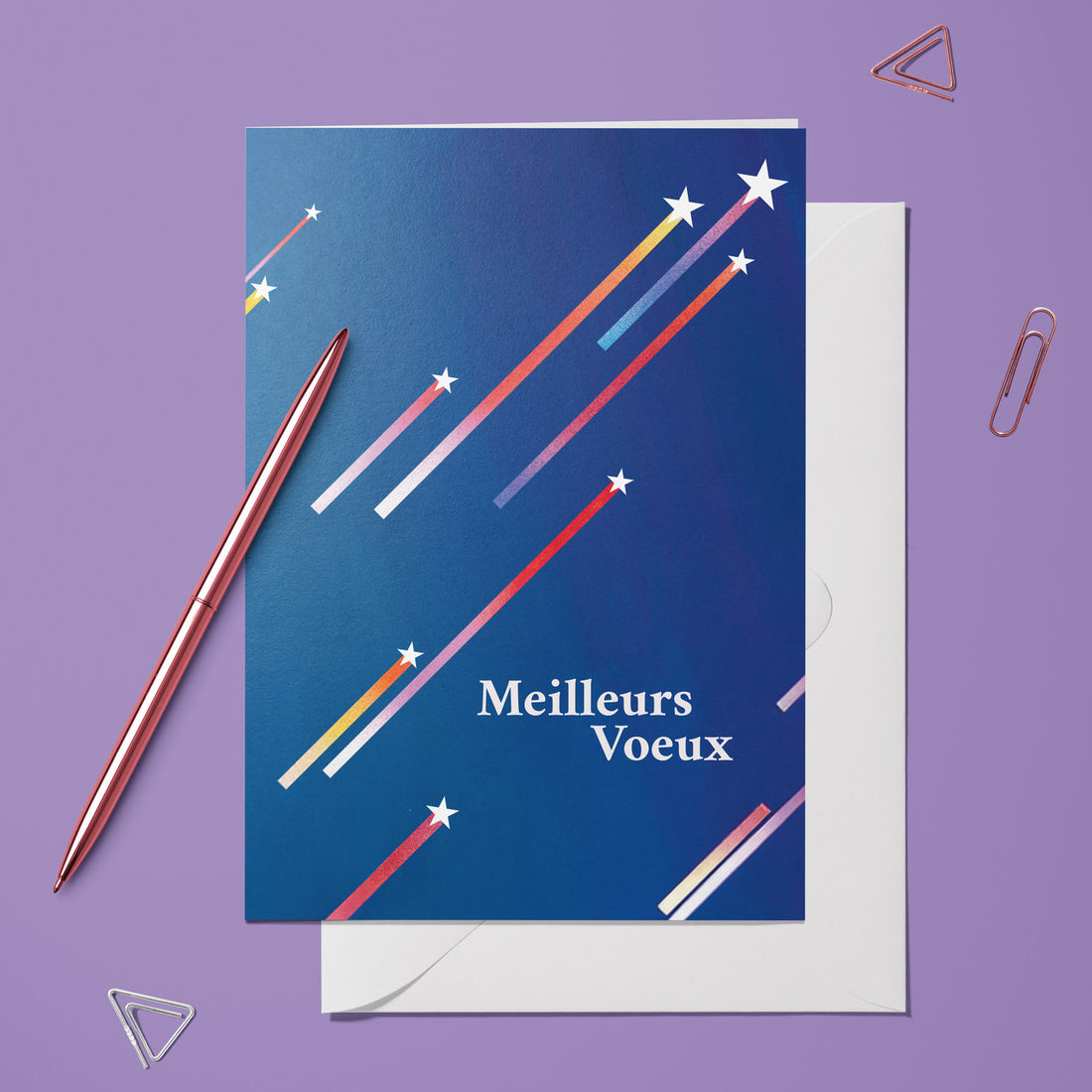 Carte pliée Meilleurs voeux posée avec un stylo doré et son enveloppe, étoiles filantes sur un ciel bleu nuit