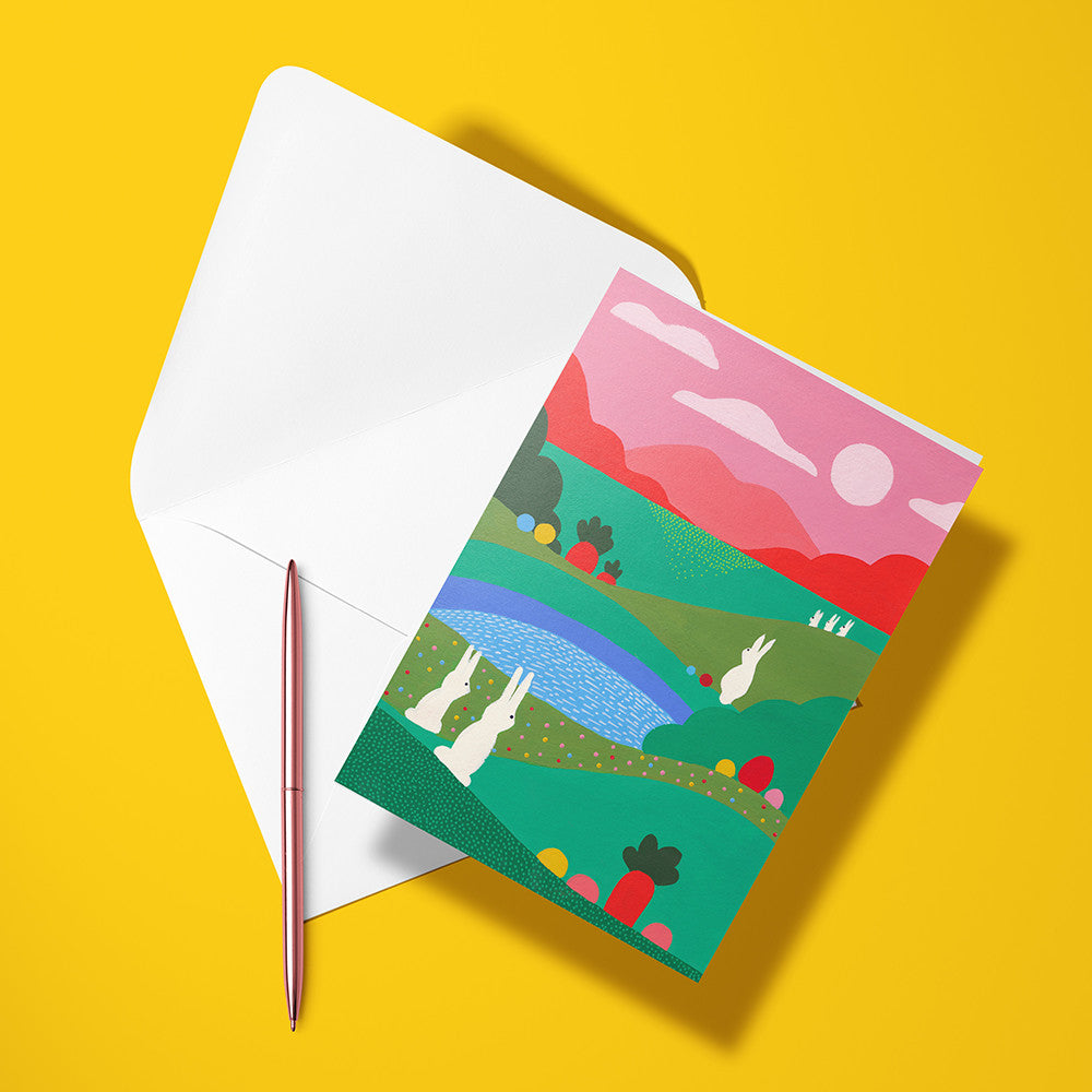 Carte pliée Pâques au jardin posée avec un stylo doré et son enveloppe, lapins blancs dans les collines vertes