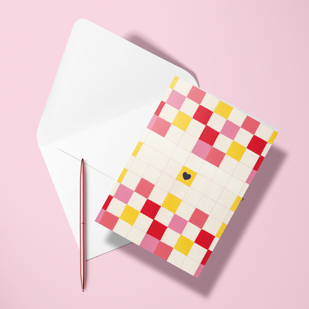 Carte pliée Damier Cœur posée avec un stylo doré et son enveloppe, damier rouge et jaune ponctué d'un cœur