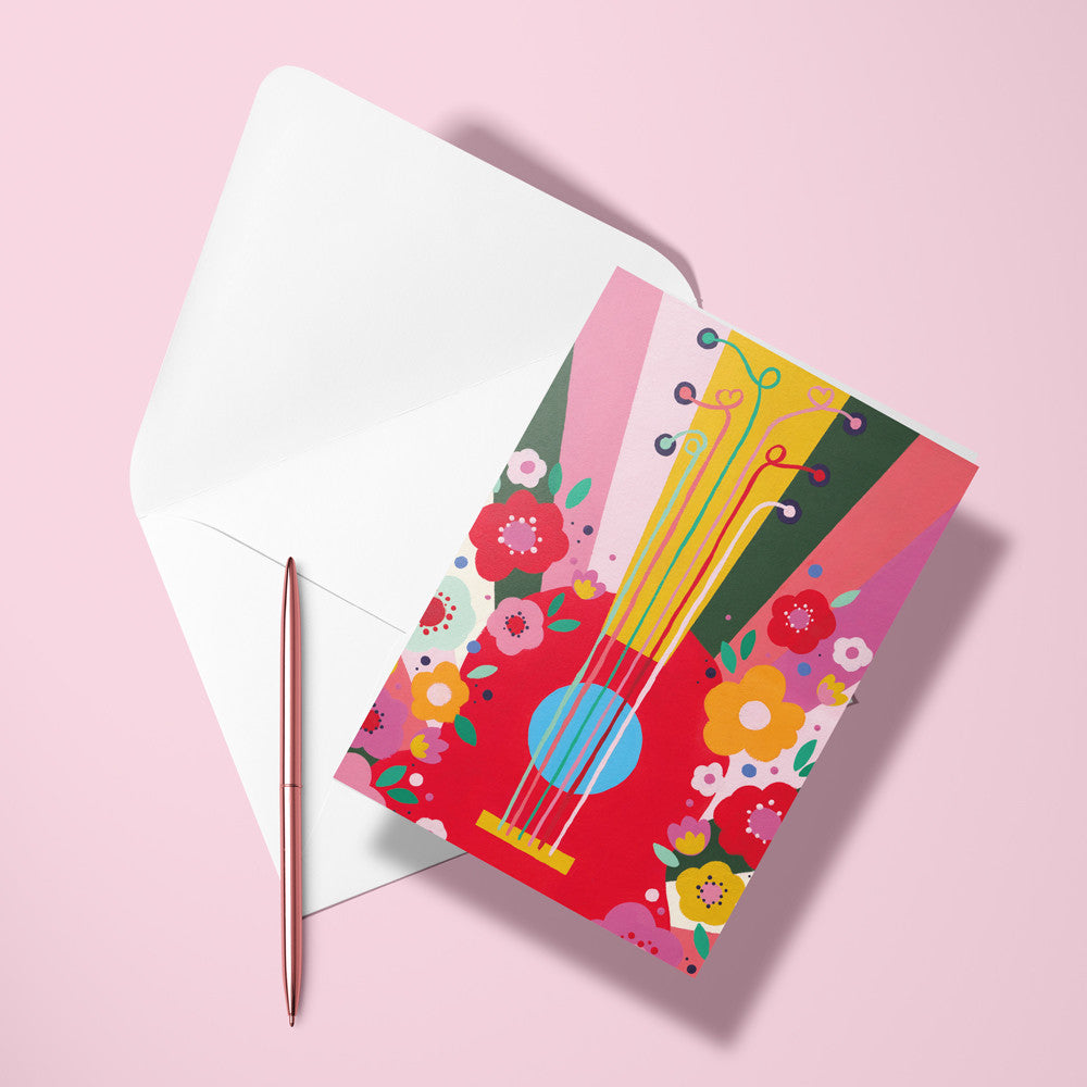 Carte pliée Guitare Flower power posée avec un stylo doré et son enveloppe sur un fond rose