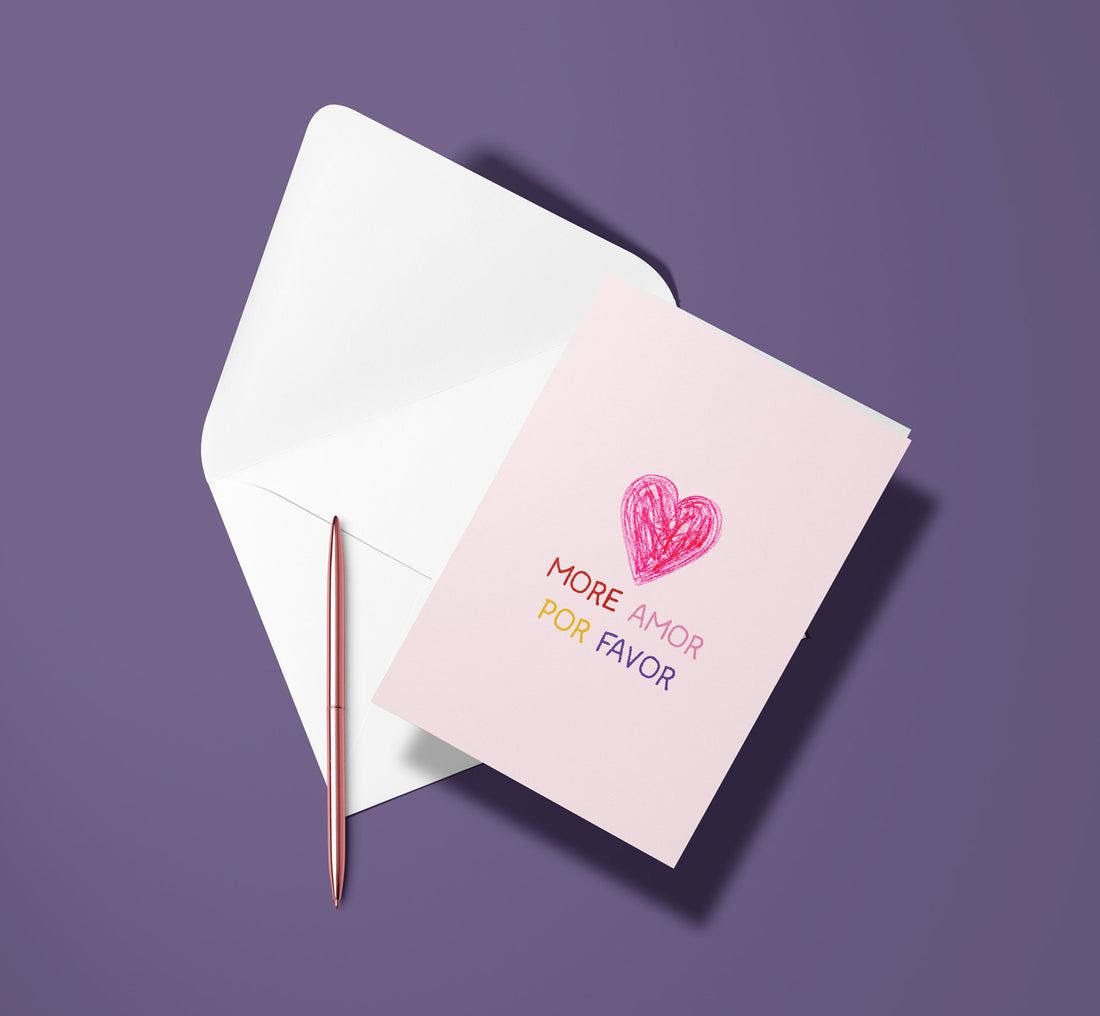 Carte pliée More Amor posée avec un stylo doré et son enveloppe, cœur rose sur fond rose pâle