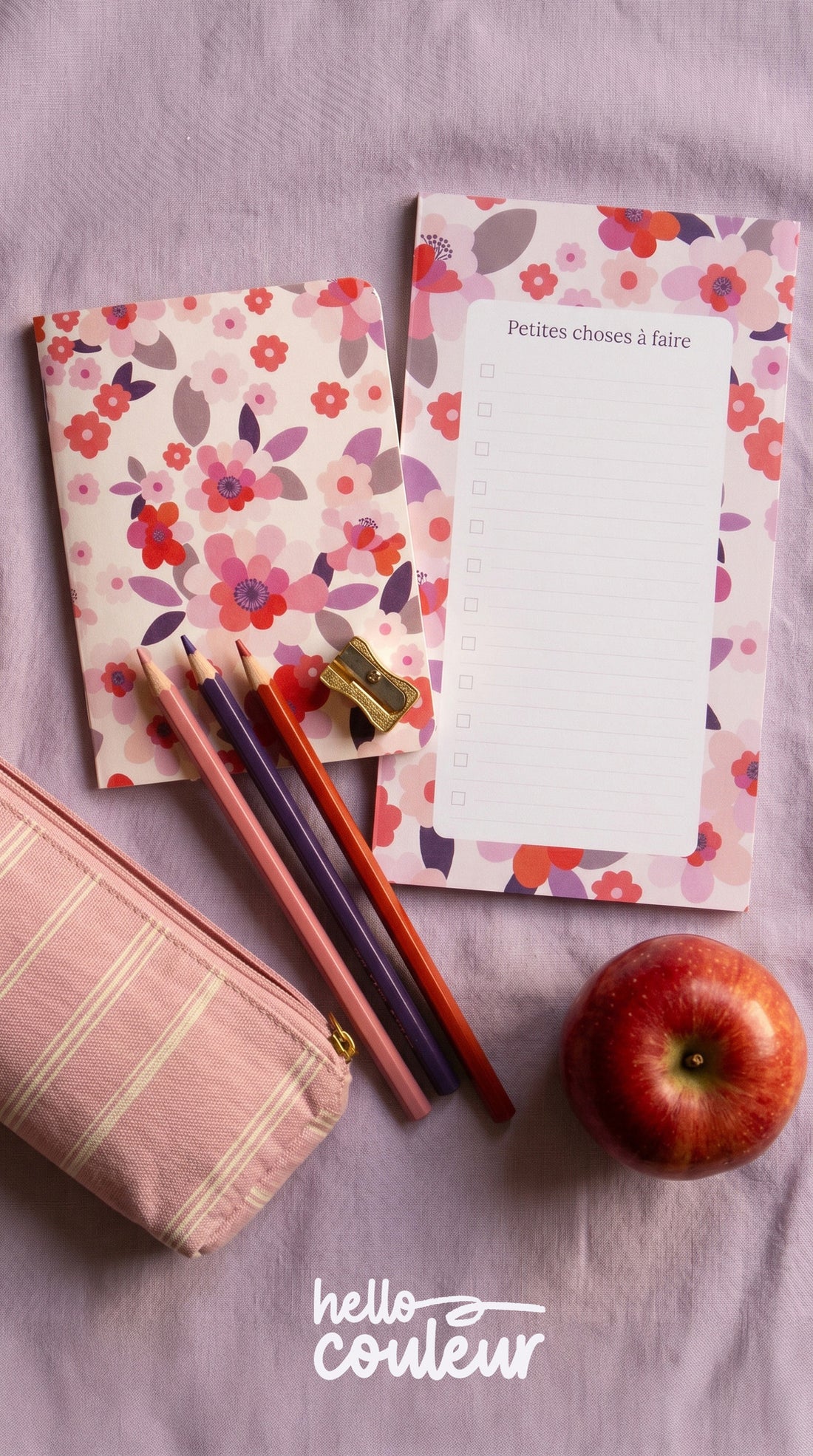 Bloc-notes Sakura présenté sur un textile rose, avec une trousse, des crayons de couleur et une pomme